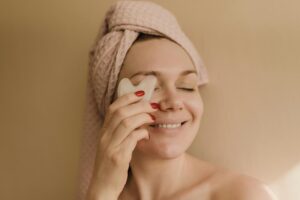 A smiling woman with a towel wrapped around her head using a gua sha tool for a facial massage.